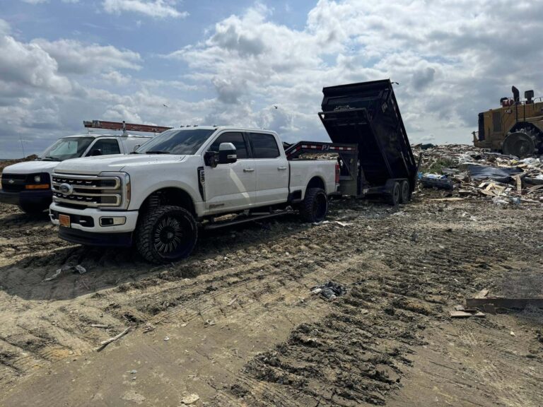 White pickup truck parked near a dumpster at a waste disposal site, with construction debris and machinery in the background, illustrating dumpster rental services and waste management solutions.