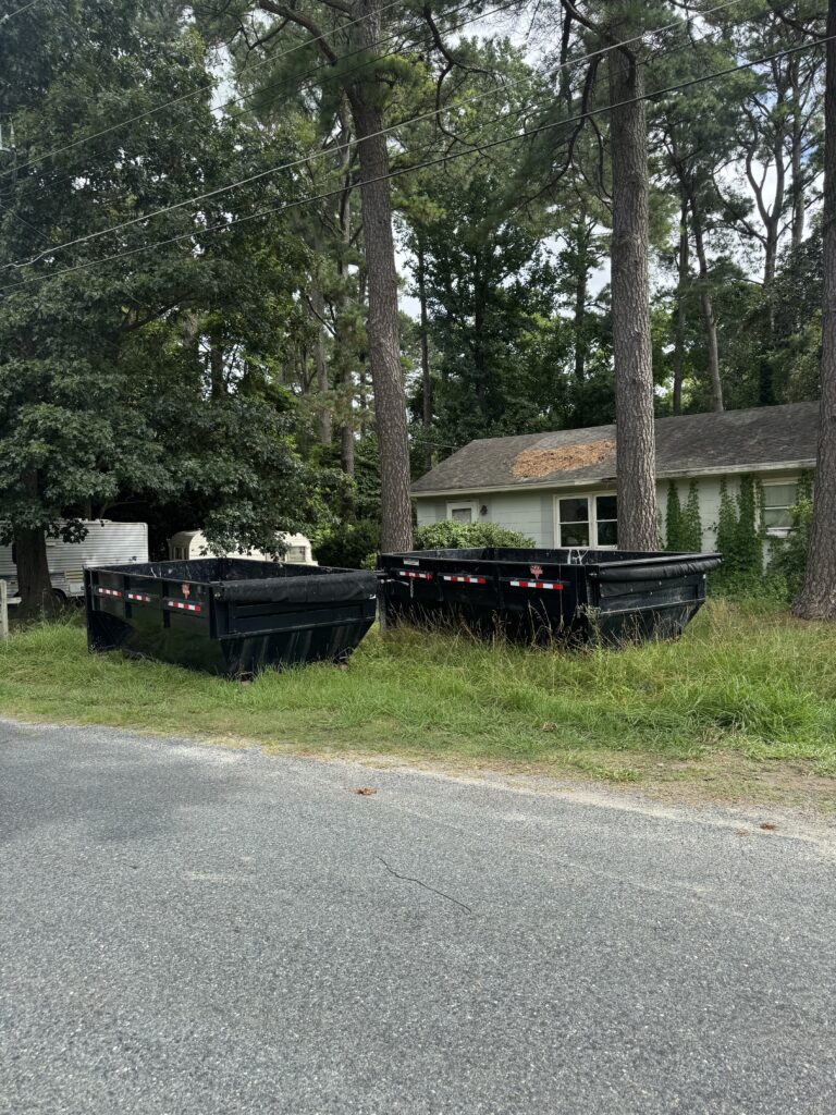 Two black dumpsters positioned on a grassy area beside a road, surrounded by trees and a residential building, illustrating waste management solutions for cleanup projects.