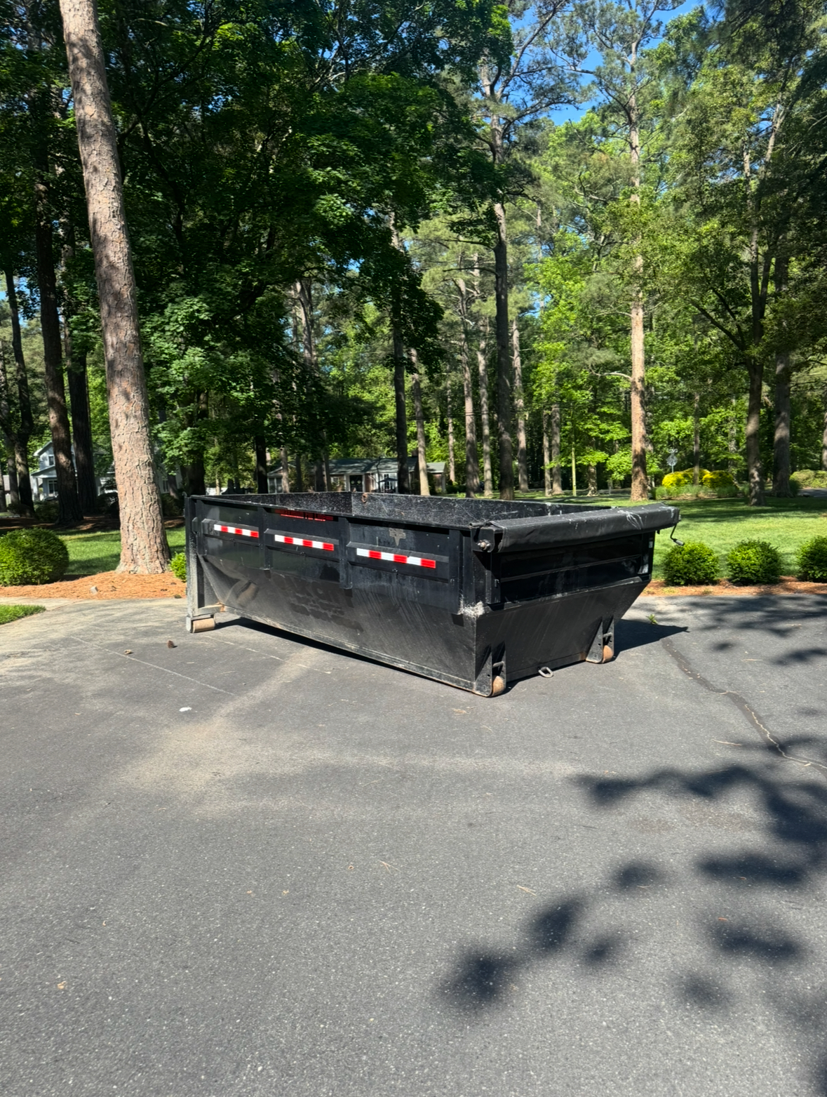 Black roll-off dumpster parked on asphalt driveway surrounded by trees, suitable for home renovation and debris disposal, emphasizing convenience for large jobs.
