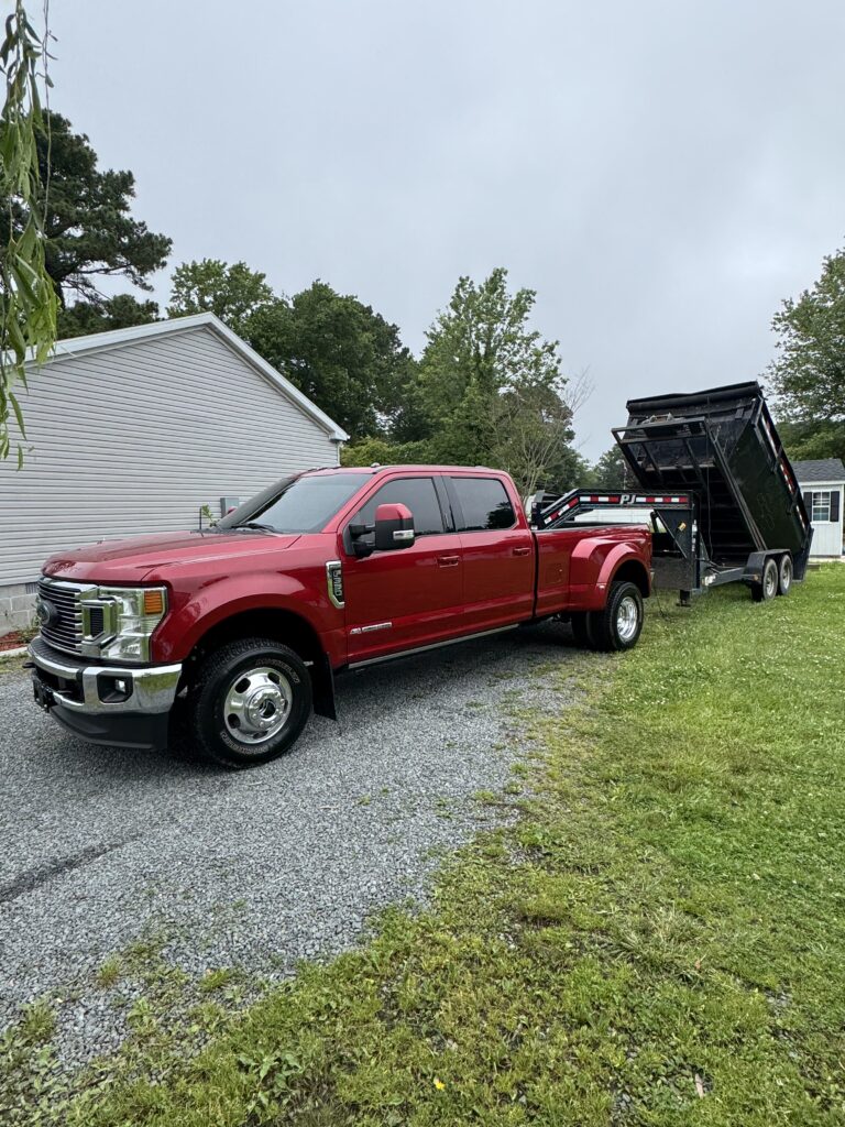 Red pickup truck parked on gravel driveway with attached black roll-off dumpster, set in a residential area in Dagsboro, DE.