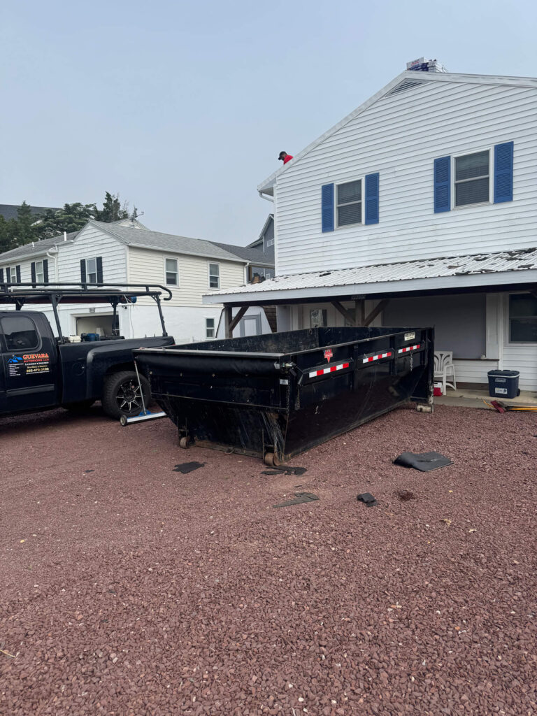 Black dumpster on gravel driveway in front of a residential home, with a worker on the roof, illustrating Home Wreckers' junk removal and construction debris services.