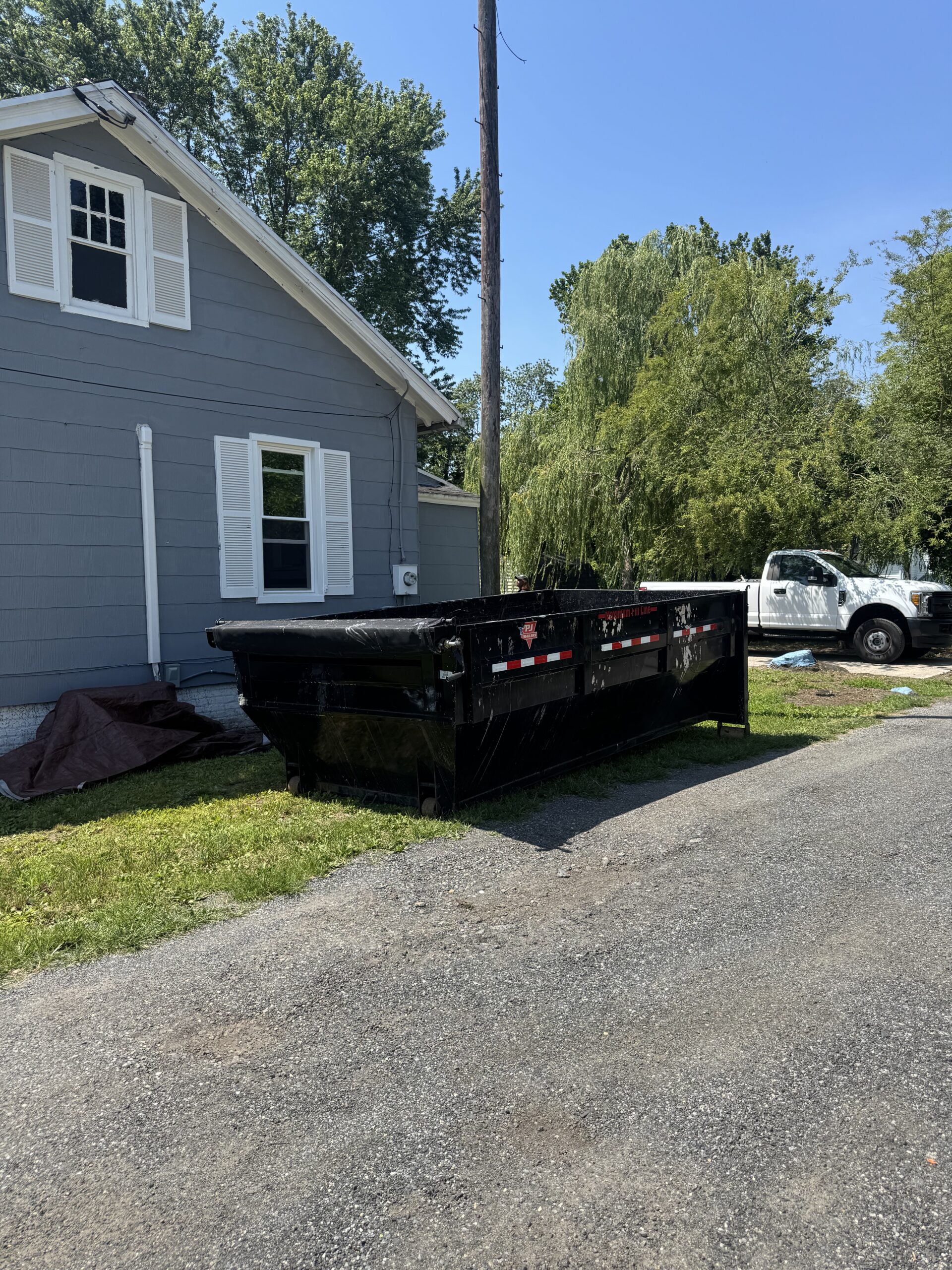 Black roll-off dumpster parked on gravel driveway beside a gray house, surrounded by greenery, illustrating dumpster rental services in Dagsboro for waste disposal needs.
