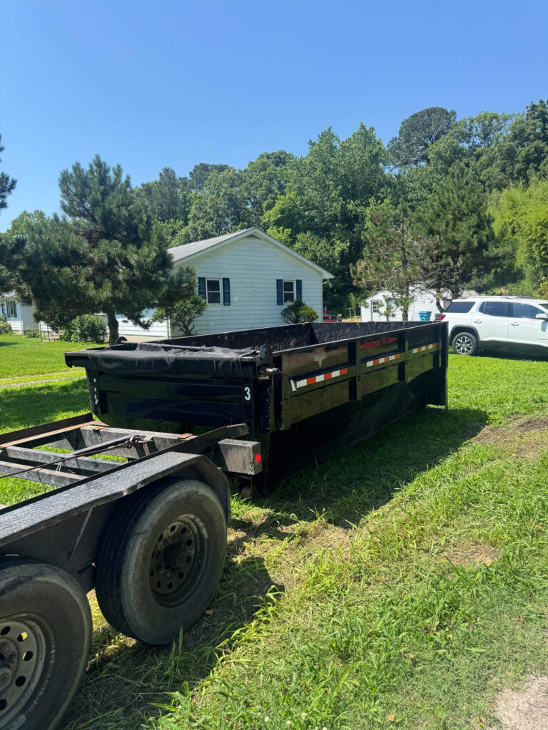 Black dumpster on grass near residential home, showcasing waste management solution for cleanup projects, surrounded by trees and parked vehicles.